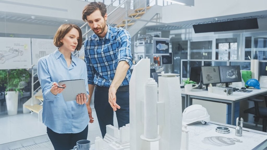 Two colleagues discussing a white architectural model, with one pointing and the other holding a tablet in a design studio.