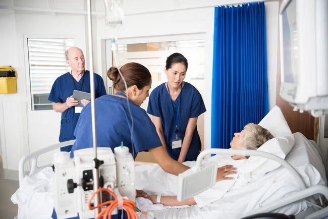 A group of healthcare professionals in blue scrubs assists an elderly patient lying in a hospital bed.