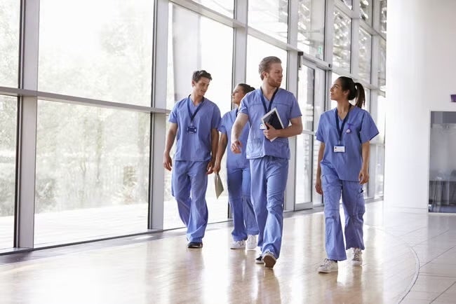 A group of medical professionals in scrubs walks through a bright hospital hallway, discussing patient care.