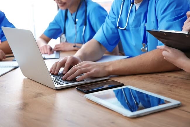 Healthcare workers in blue scrubs work together using a laptop and tablets at a wooden table.