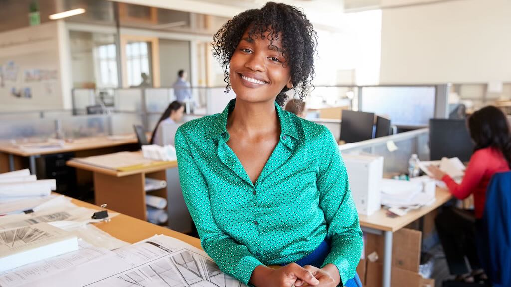A smiling woman wearing a green shirt, sitting at a desk surrounded by papers in an open office environment.