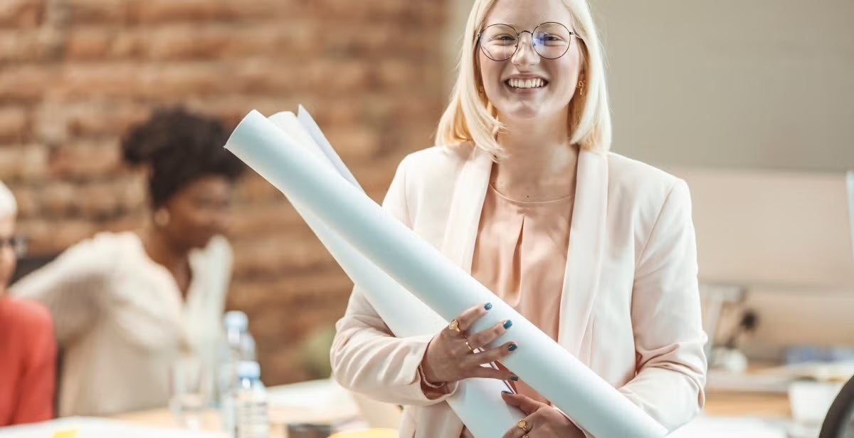 A cheerful woman holding rolled-up blueprints in an office setting, with a colleague blurred in the background.