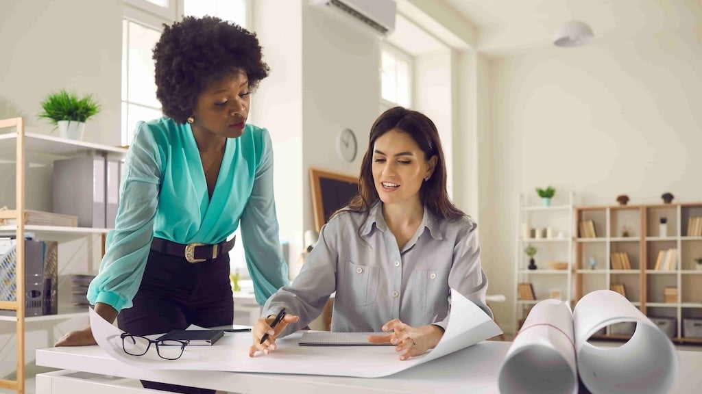 Two women, one standing and one seated, discuss a project while reviewing documents at a desk.