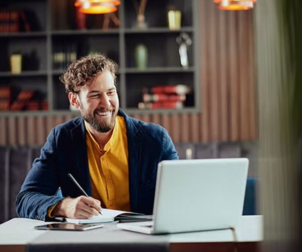 Man in a blue blazer, writing and working on a laptop in a library or office space with shelves in the background.