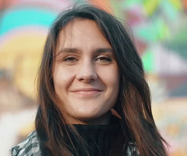 Woman standing outdoors with a slight smile, colorful background with a blurred mural.