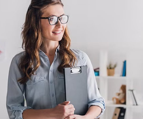 Smiling woman in glasses, holding a clipboard and standing in an office space.