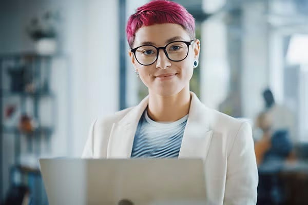 A woman in glasses confidently smiles while she holds her laptop in the office.