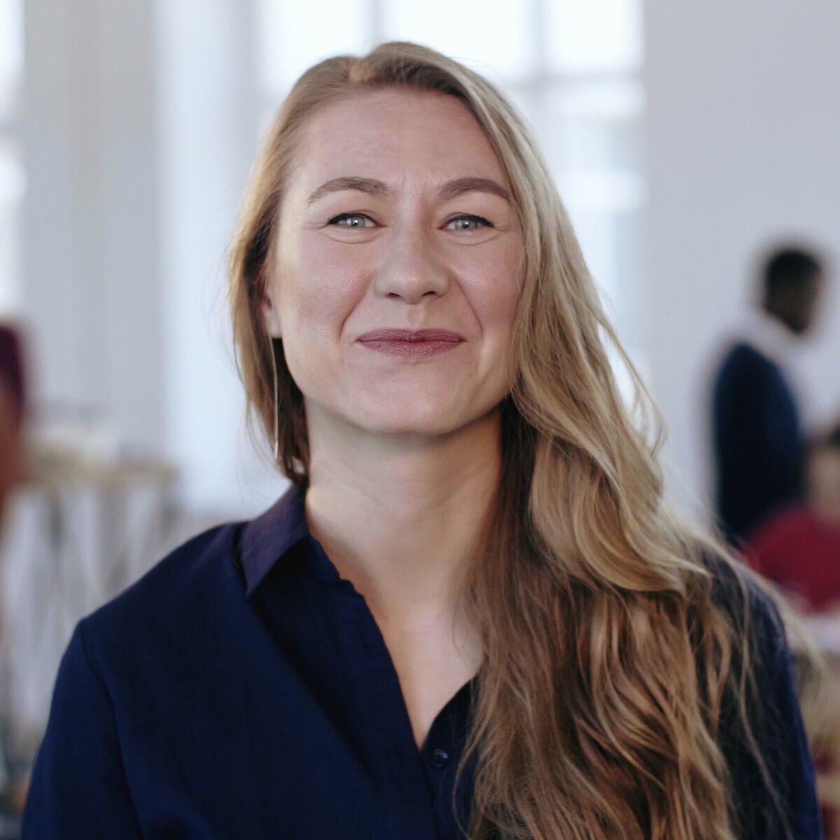 A smiling woman with long blonde hair wearing a navy blue shirt in a bright, modern office.
